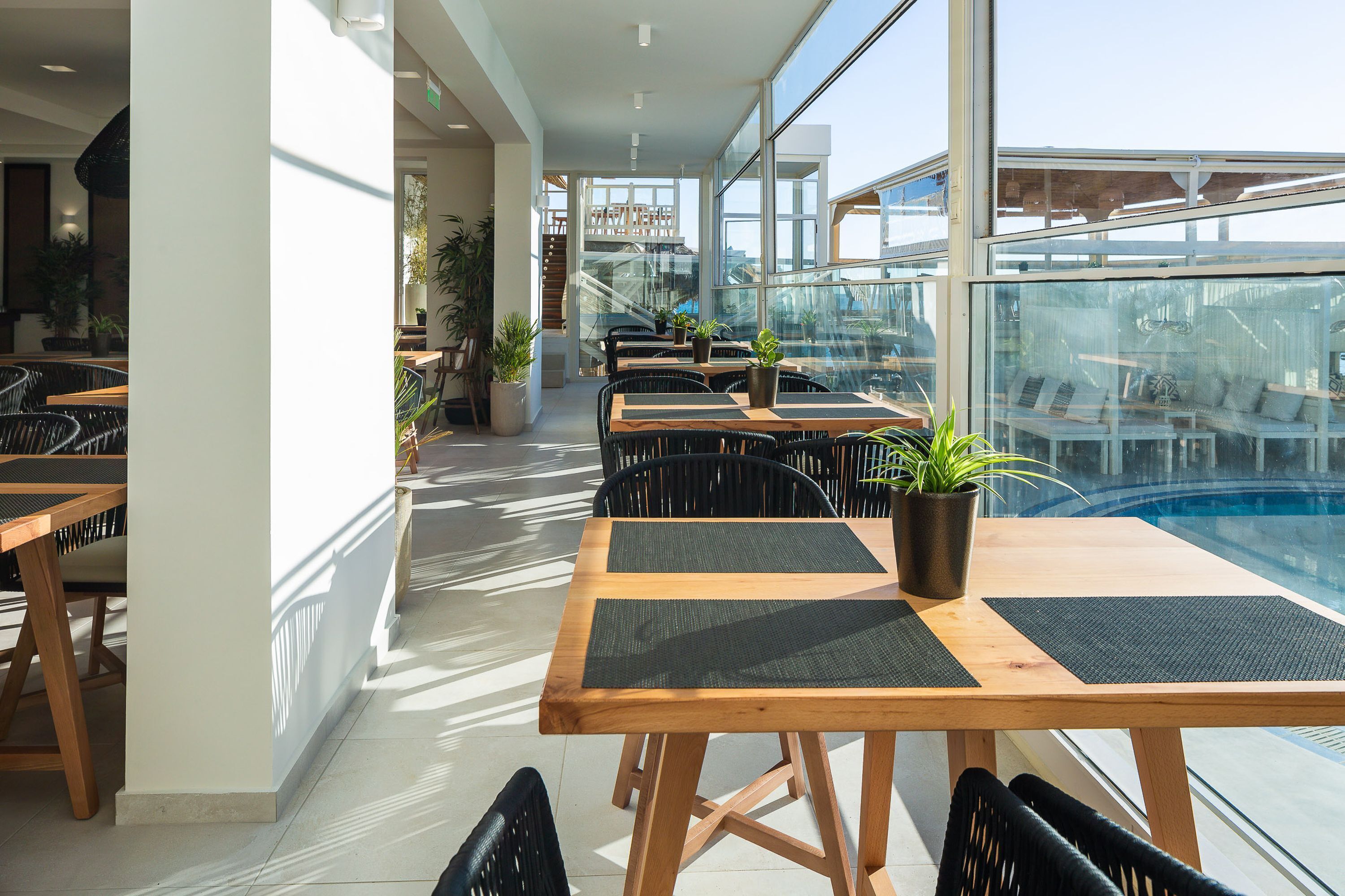 Un restaurante en la azotea con mesas y sillas de madera, plantas en macetas y vistas a la piscina.