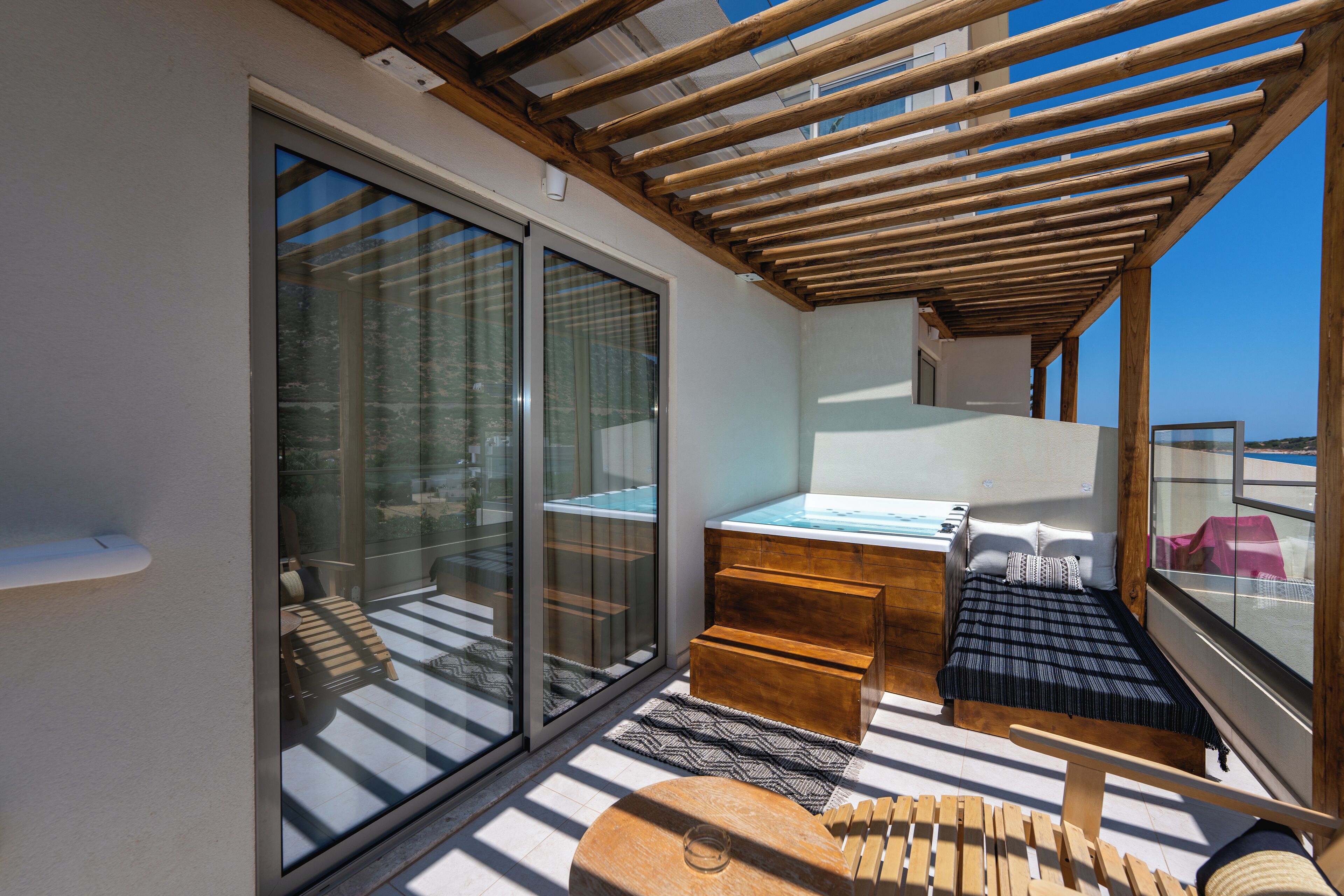 A balcony with a wooden pergola, a glass door leading to a seating area, and a small bed with a blue bedspread.