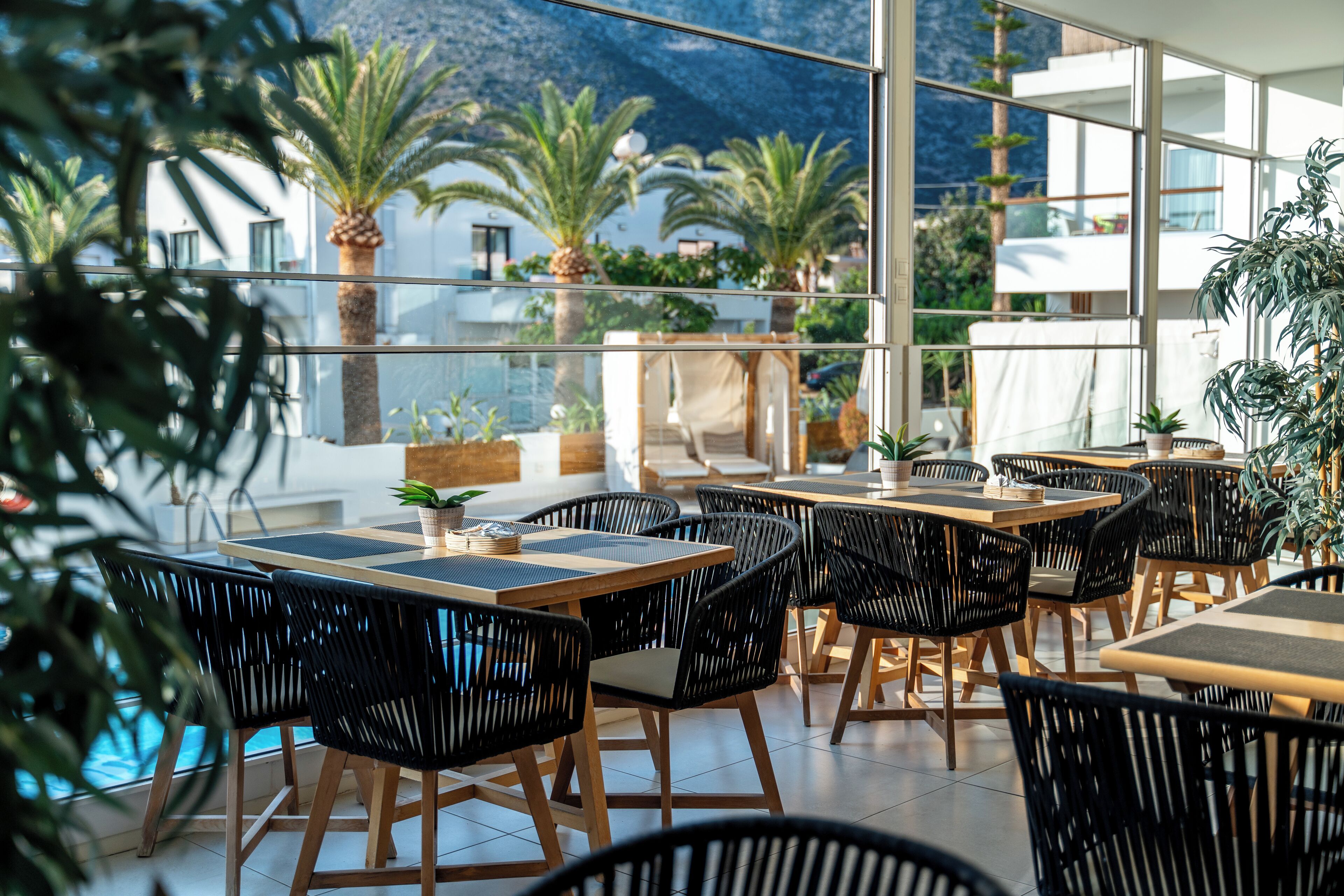 A restaurant with outdoor seating, black wicker chairs, wooden tables, and potted plants.