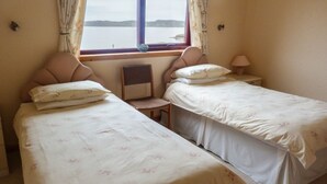 A bedroom with two single beds, a chair, a nightstand, a lamp, and a window with curtains. - 2 Bayview Bungalow (Achnasheen)