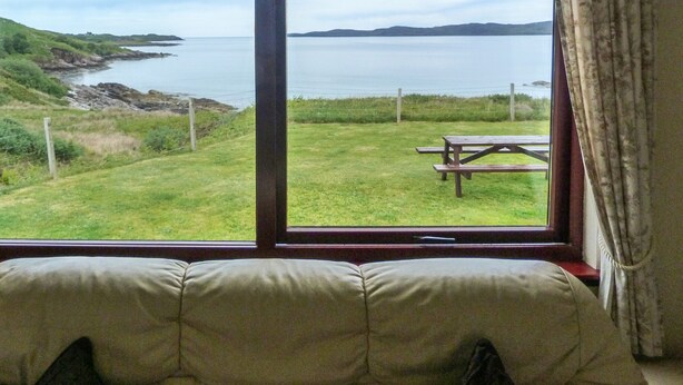 A room with a large window overlooking a grassy area with a picnic table. - 2 Bayview Bungalow (Achnasheen)