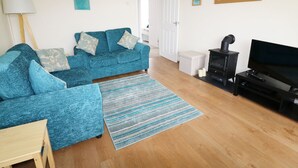 A living room with a blue sofa set, a wooden coffee table, a striped rug, a black TV stand, and a wood-burning stove. - Anneddle (Holyhead)