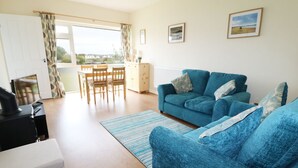 A living room with a blue sofa, a dining table with chairs, and a view of the outdoors through a large window. - Anneddle (Holyhead)
