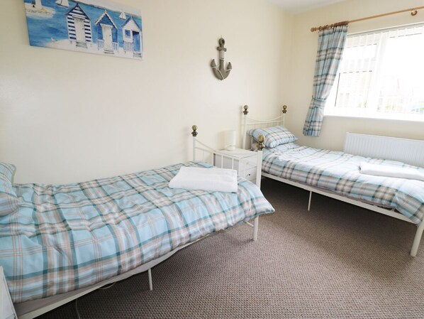Un dormitorio con dos camas individuales, una mesita de noche, una lámpara, un cuadro con cabañas de playa y una ventana con cortinas.