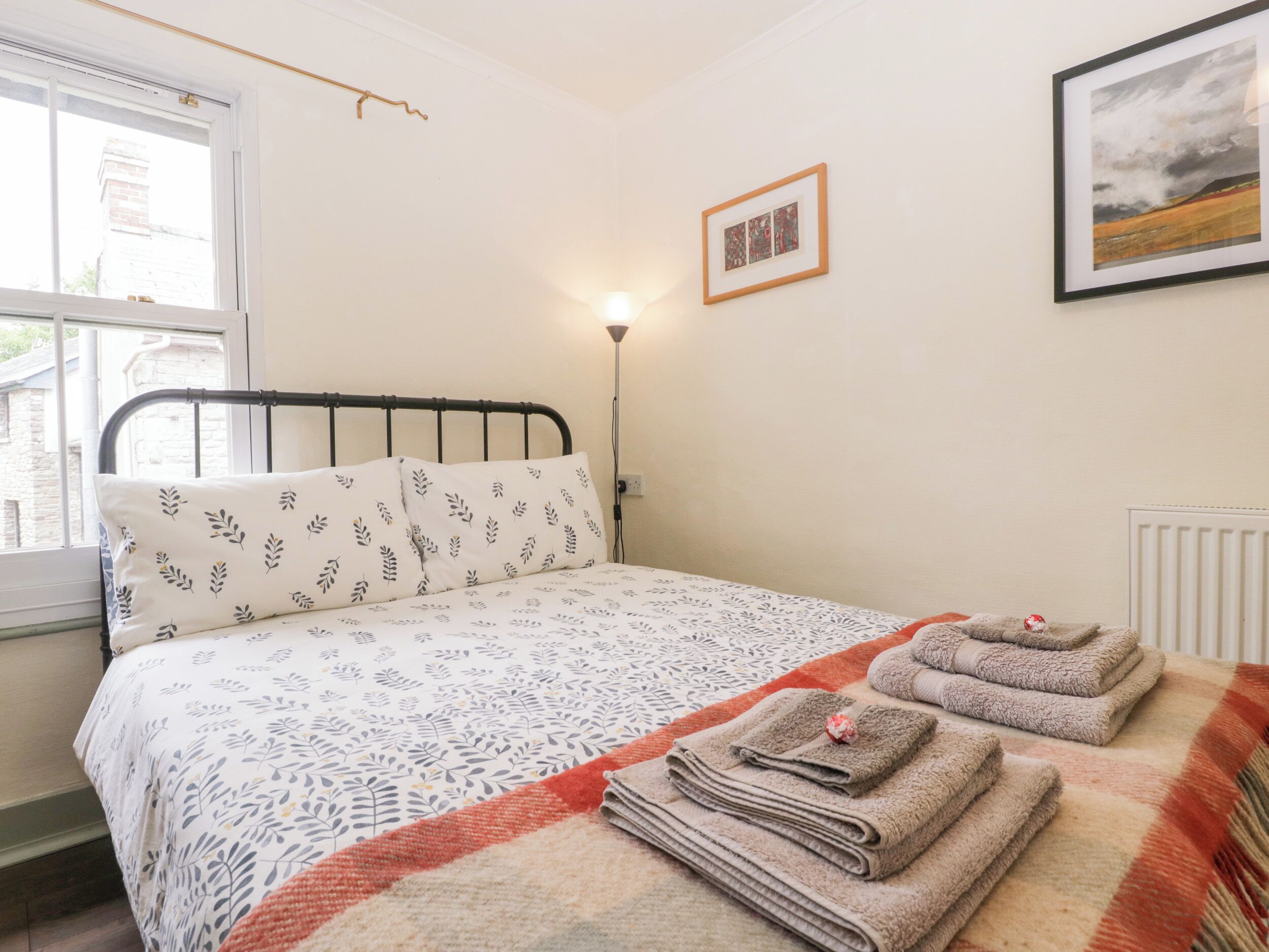 Un dormitorio con cama, almohadas, una lámpara de noche, un cuadro enmarcado y una ventana.