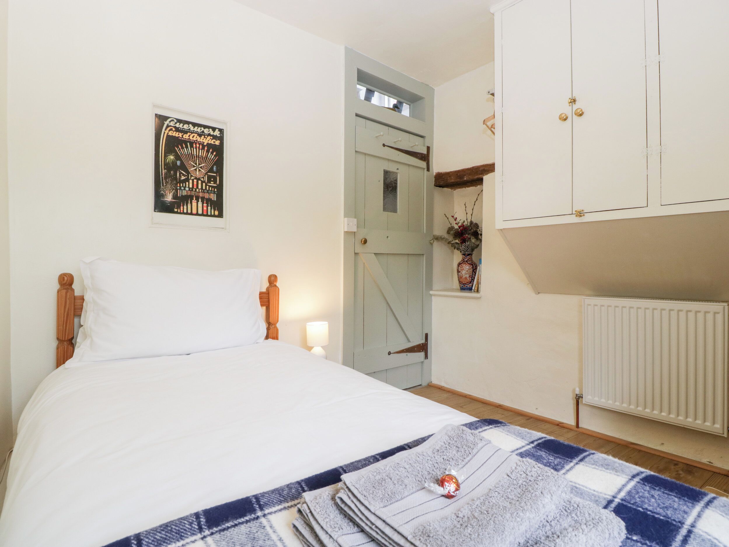A bedroom with a bed, a nightstand, a wall-mounted lamp, a door leading to another room, and a radiator.