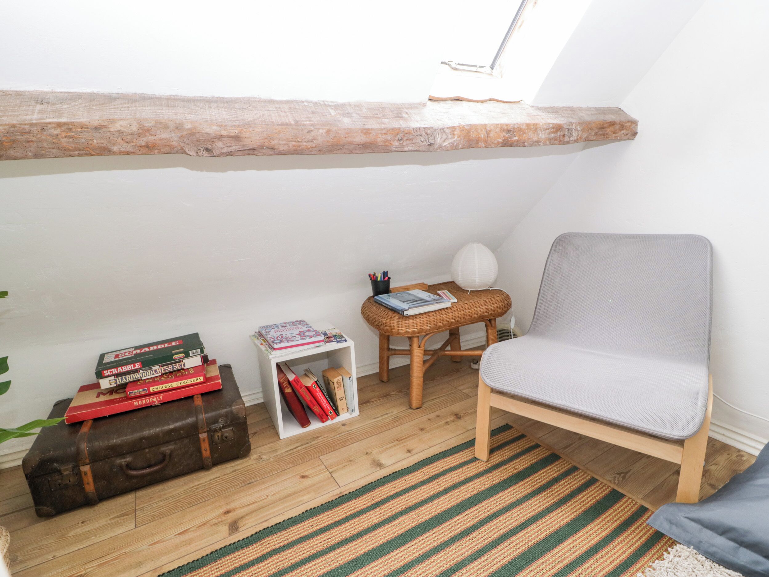 A cozy attic room with a sloped ceiling, a wicker chair, a small table with books and a lamp, and a suitcase on the floor.