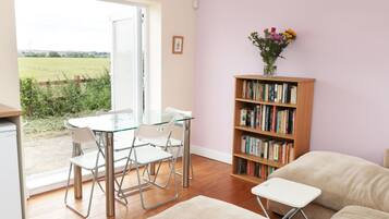 A cozy living room with a glass table and chairs, a bookshelf, and a view of a green field through a sliding door.