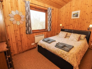 Ein Schlafzimmer mit Holzwand, einem Bett, einem Heizkörper, einem Fenster mit Vorhängen, einer Wanduhr und einem gerahmten Bild.