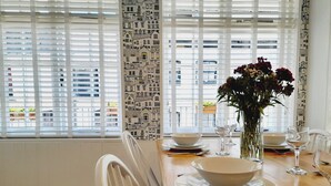 A dining area with a wooden table set for two, white chairs, a vase with flowers, and a view of buildings through large windows with blinds. - Crab Cottage (Ilfracombe)