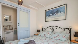 Een slaapkamer met een bed, nachtkastje, lamp, een ingelijst schilderij aan de muur, en een badkamer met wastafel en spiegel.