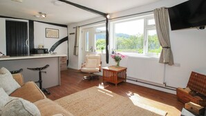 Een gezellige woonkamer met een houten vloer, een bank, een fauteuil, een bijzettafeltje met een vaas, en een televisie aan de muur.