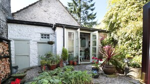 A white building with a stone roof, a garden with various potted plants, and a pile of firewood. - Mill Bridge Cottage (Hope Valley)