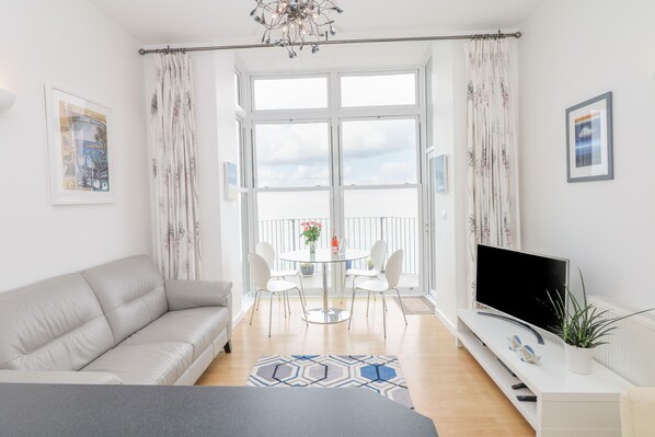 A modern living room with a sofa, a glass table with chairs, a TV on a stand, and a balcony with curtains. - Oceans Side (Ilfracombe)