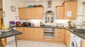 A modern kitchen with wooden cabinets, a built-in oven, microwave, and dishwasher. There is a sink, a fruit bowl, and various kitchen utensils on the counter. - 12 West End Point (Pwllheli)