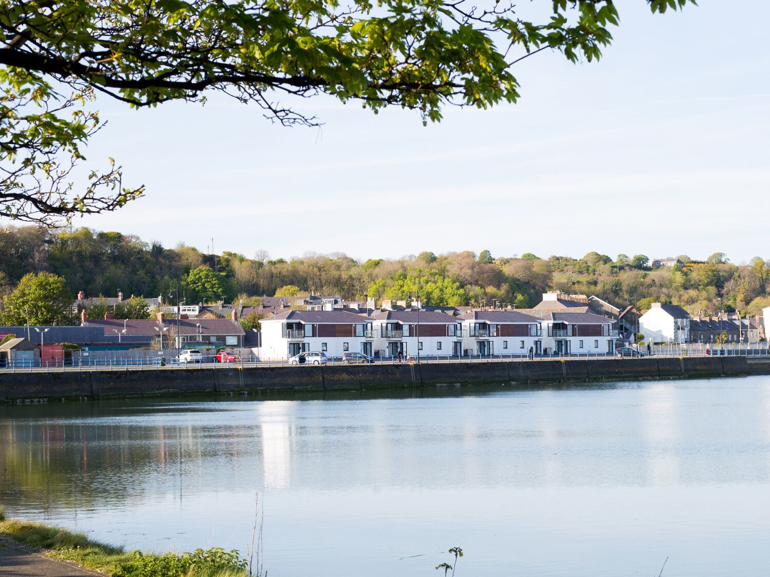 A waterfront view with modern buildings, a calm river, and a tree-lined hill in the background.