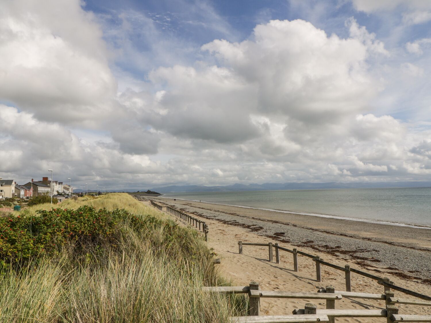 A coastal beach with a wooden walkway, grassy dunes, and houses in the background.