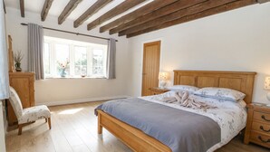 A bedroom with a wooden bed, a nightstand, a chair, a window with curtains, and a wooden ceiling. - Glan Felin (Pwllheli)