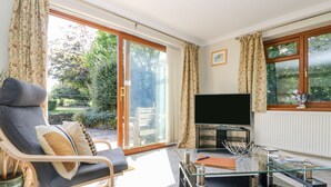 A modern living room with a glass coffee table, a comfortable lounge chair, a flat-screen TV, and a sliding glass door leading to a garden. - Silverwell Forge (Truro)