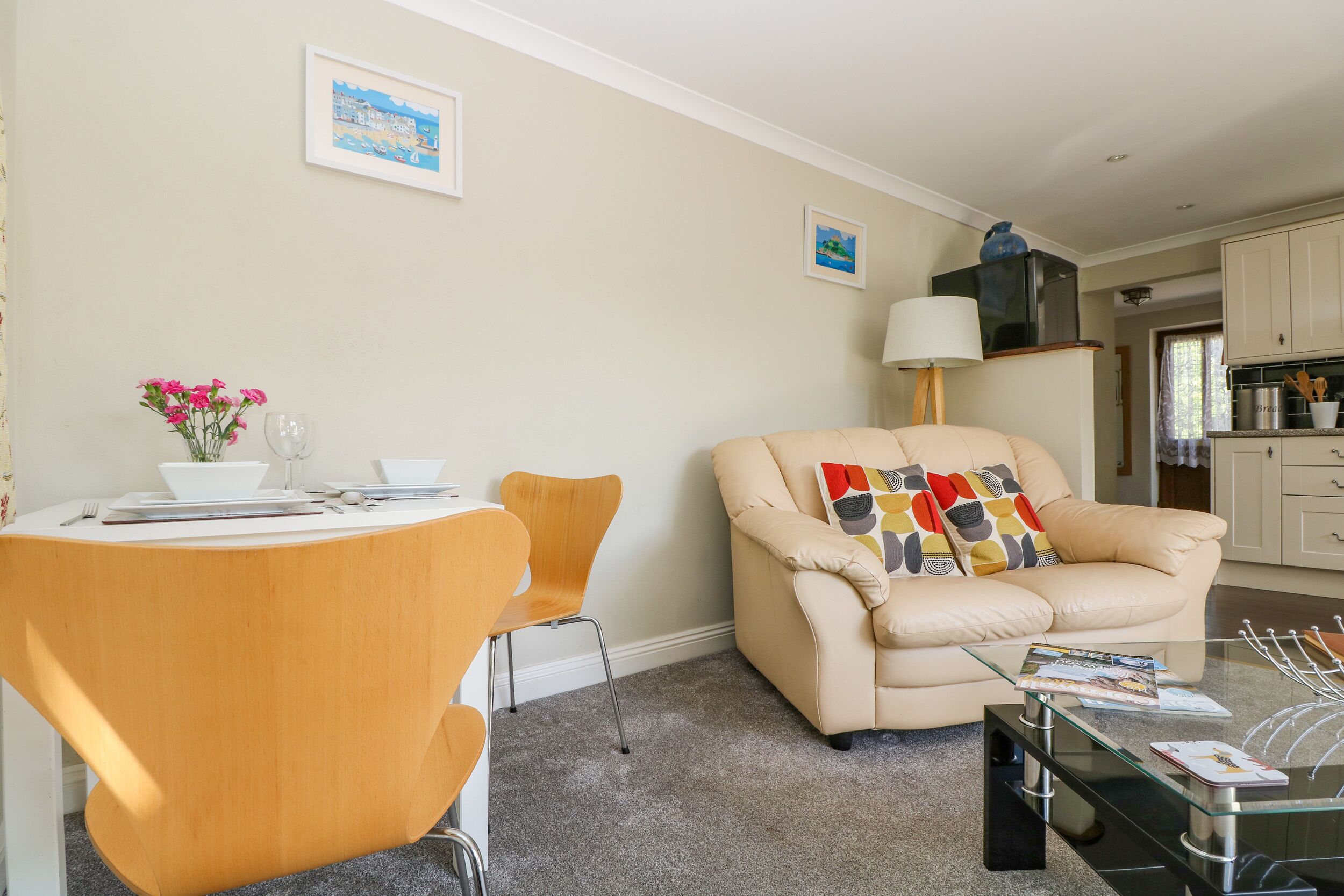 A living room with a beige sofa, a glass coffee table, a dining table with chairs, and a kitchen area in the background.