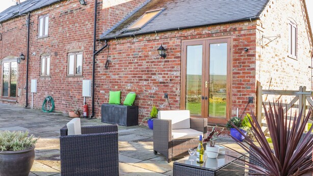 A brick building with a patio area featuring wicker furniture, a glass table, and potted plants. - Clun Forest Cottage (Much Wenlock)