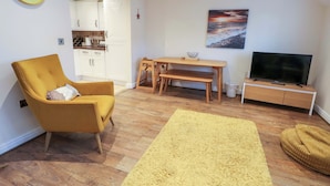 A living room with a yellow armchair, a wooden dining table, a TV on a stand, and a painting of a sunset on the wall.
