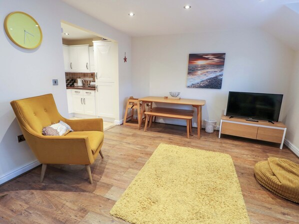 Un salon avec un fauteuil jaune, une table à manger en bois avec des bancs, un téléviseur posé sur un meuble et un tableau représentant un coucher de soleil accroché au mur.