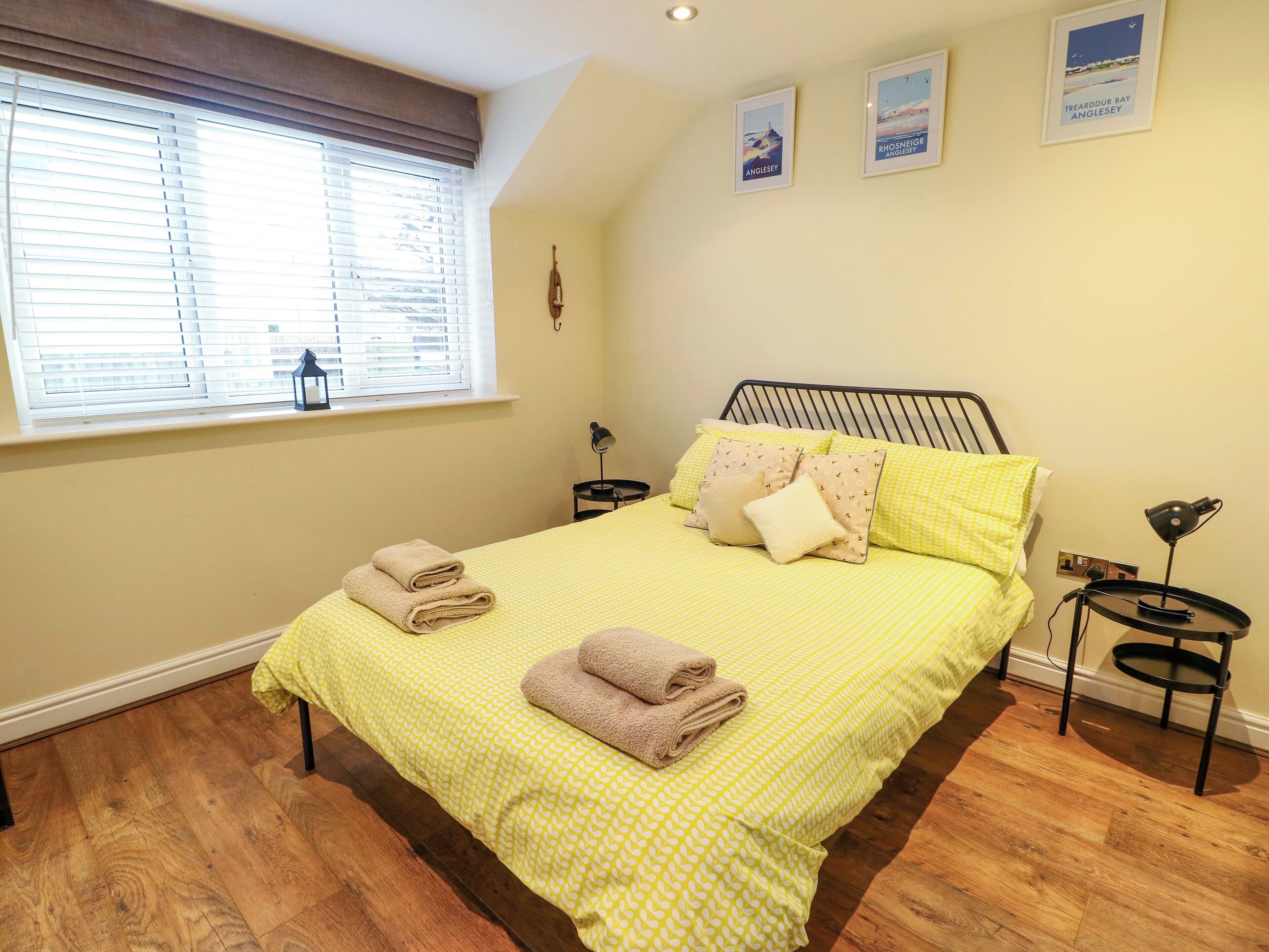 A bedroom with a bed, pillows, a bedside table, a lamp, and a window with blinds.
