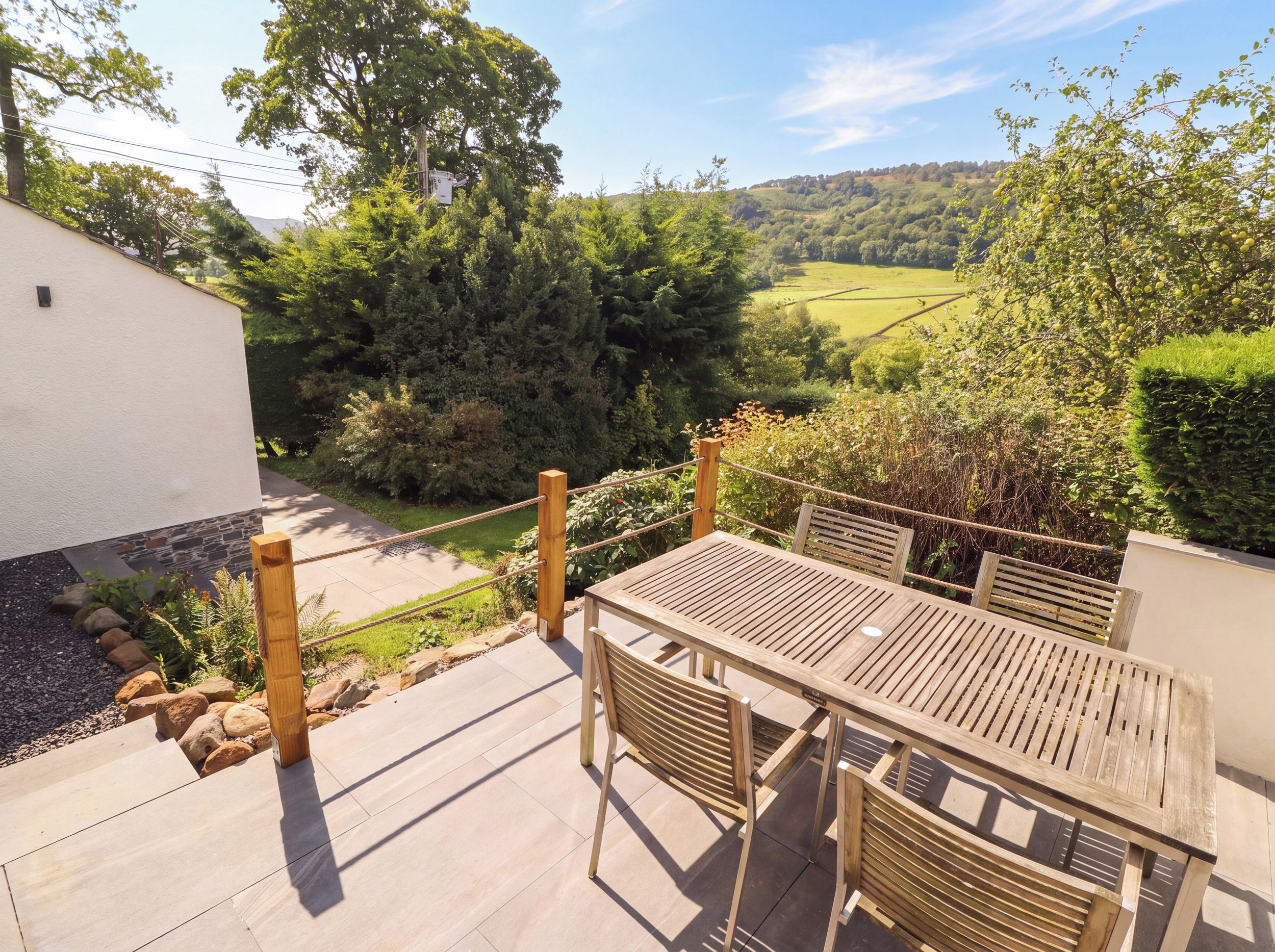 An outdoor patio with a table and chairs, surrounded by a garden and a view of a green landscape.