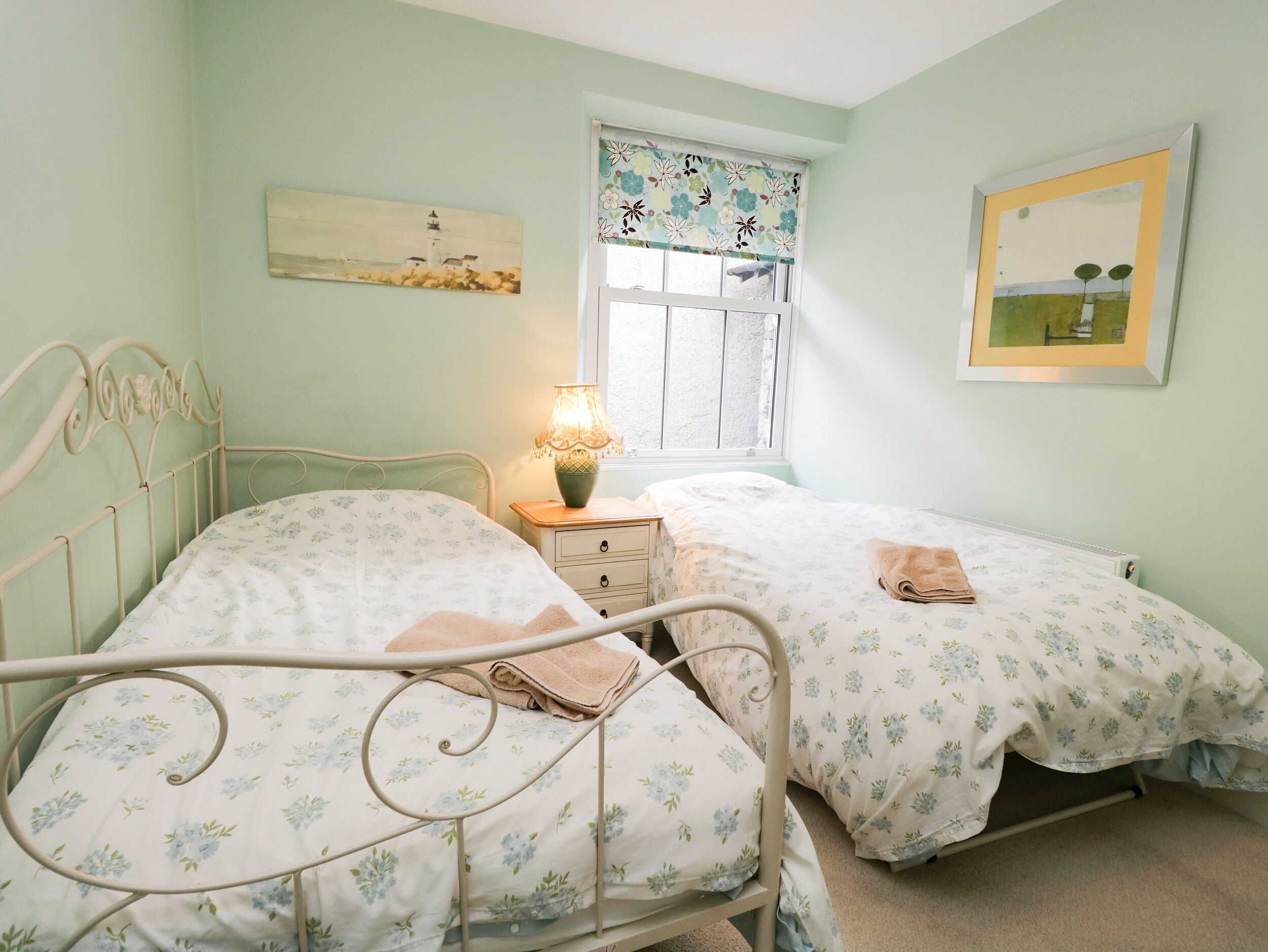 A bedroom with two single beds, a nightstand with a lamp, a window with floral curtains, and a painting on the wall.