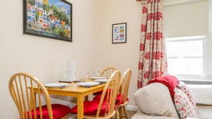 A dining area with a wooden table set for four, surrounded by wooden chairs with red cushions. A framed painting of colorful buildings is on the wall, and a window with floral curtains is visible. - Castlerigg (Keswick)