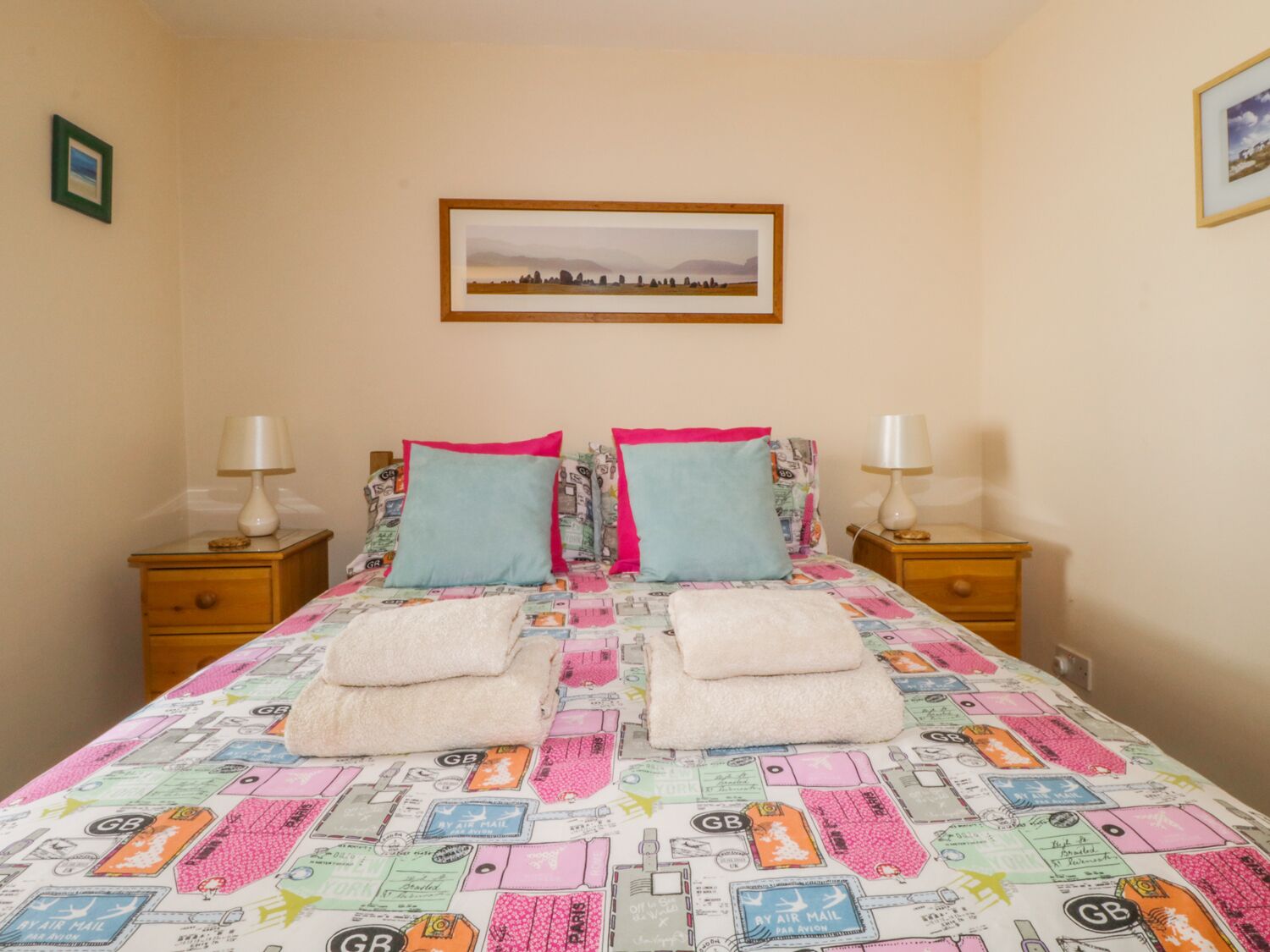 A bedroom with a bed covered in a colorful blanket, two pillows, two bedside tables with lamps, and a framed picture on the wall.