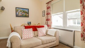 A cozy living room with a beige sofa, red and white polka dot cushions, a red throw blanket, and a red rug. A framed painting of a colorful village is on the wall, and a window with floral curtains is visible. - Castlerigg (Keswick)