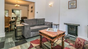 A living room with a grey sofa, a wooden coffee table, a fireplace, and a dining area with chairs and a table. - Fountain Cottage (Keswick)