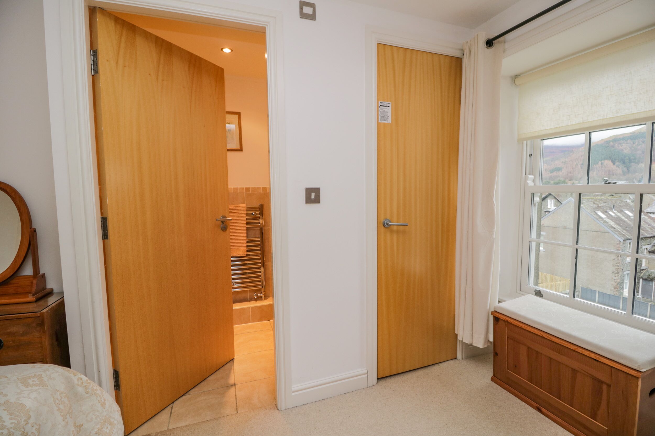 A bedroom with a wooden door, a window with a view, and a bedside table.
