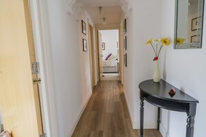 Um corredor com piso de madeira, um console preto com um vaso de flores e um espelho na parede.