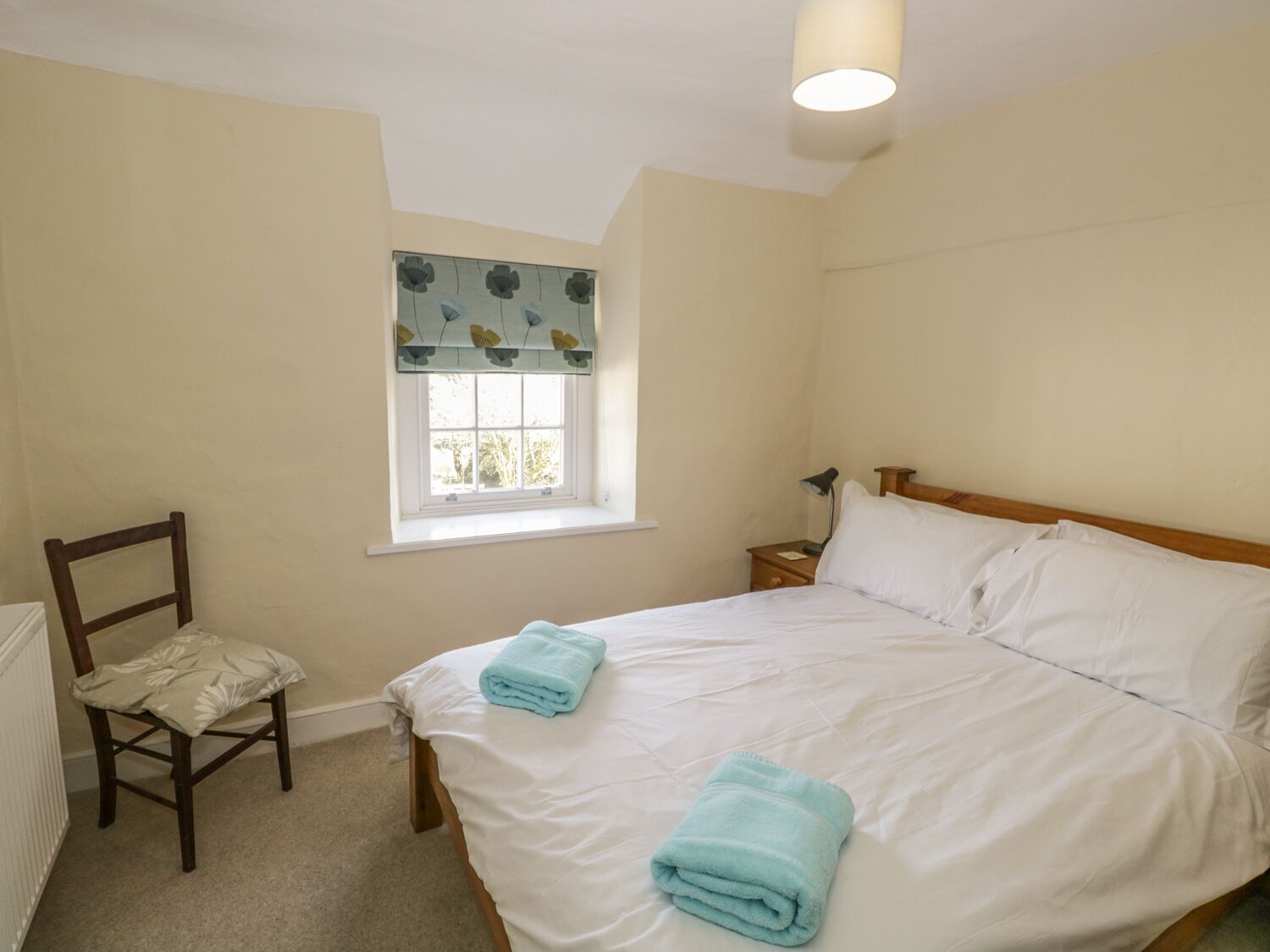 Une chambre à coucher avec un lit, une chaise, une table de chevet, une lampe et une fenêtre avec un rideau.
