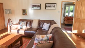 A living room with a brown leather sofa, wooden coffee table, and a floor lamp. There are two framed pictures on the wall and a door leading to another room. - Foresters Cottage (Ulverston)
