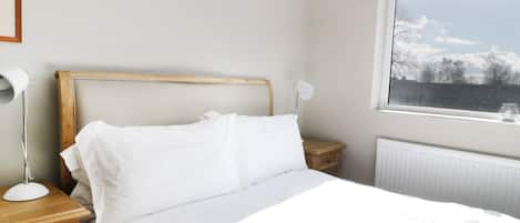A neatly made bed with white linens, a wooden headboard, a bedside table with a lamp, and a window with a view of trees and sky.