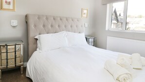 Un lit bien fait, avec du linge de lit blanc, une tête de lit capitonnée et une fenêtre donnant sur des immeubles.