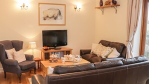 A living room with a dark brown leather sofa, a grey armchair, a wooden coffee table with pastries, a flat-screen TV, and a framed picture on the wall. - Ryelands Cottage (Much Wenlock)