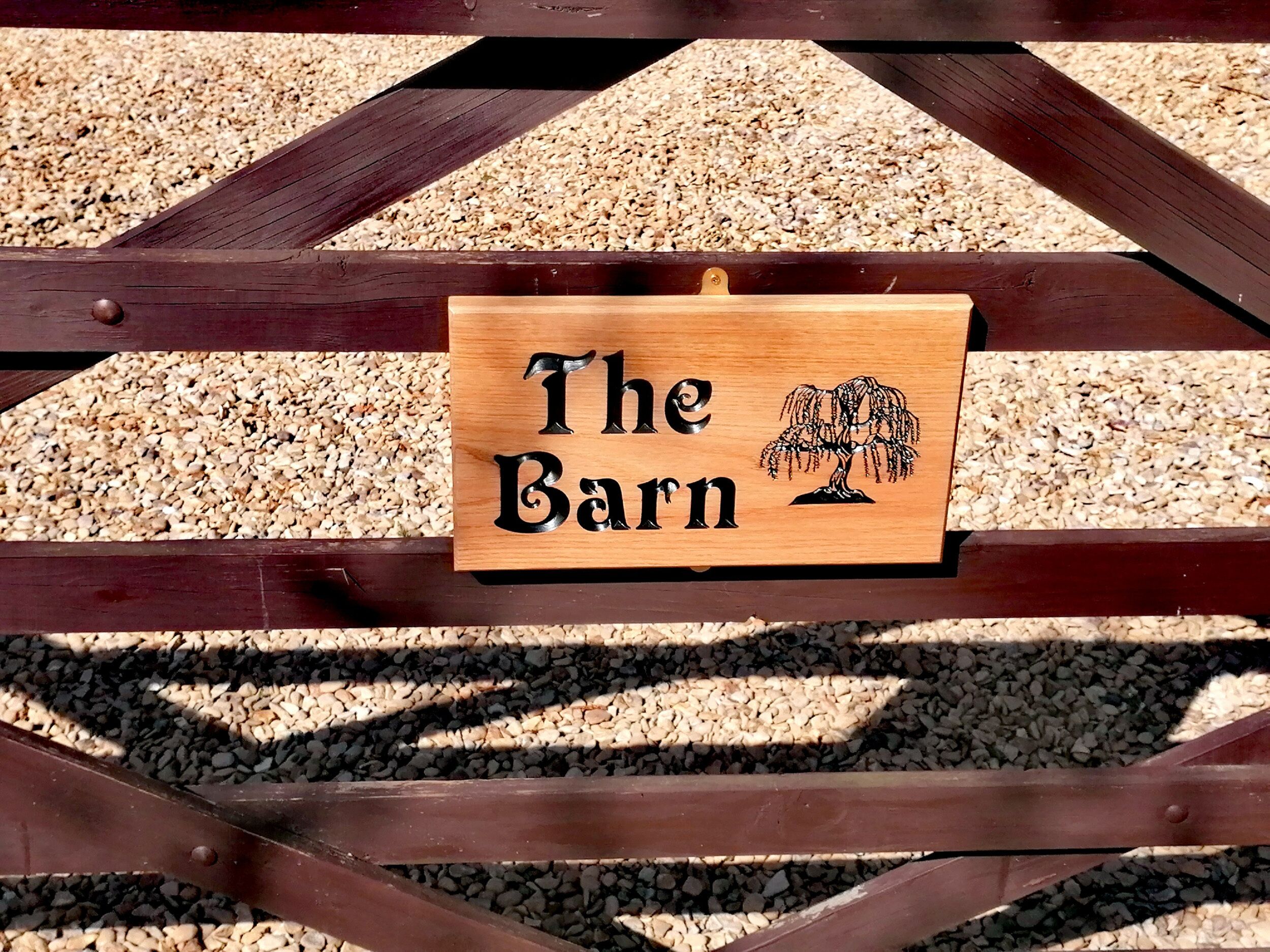Een houten bord met de tekst 'The Barn' en een illustratie van een boom, bevestigd aan een houten constructie, met grind op de achtergrond.