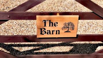 Een houten bord met de tekst 'The Barn' en een illustratie van een boom, bevestigd aan een houten constructie, met grind op de achtergrond.
