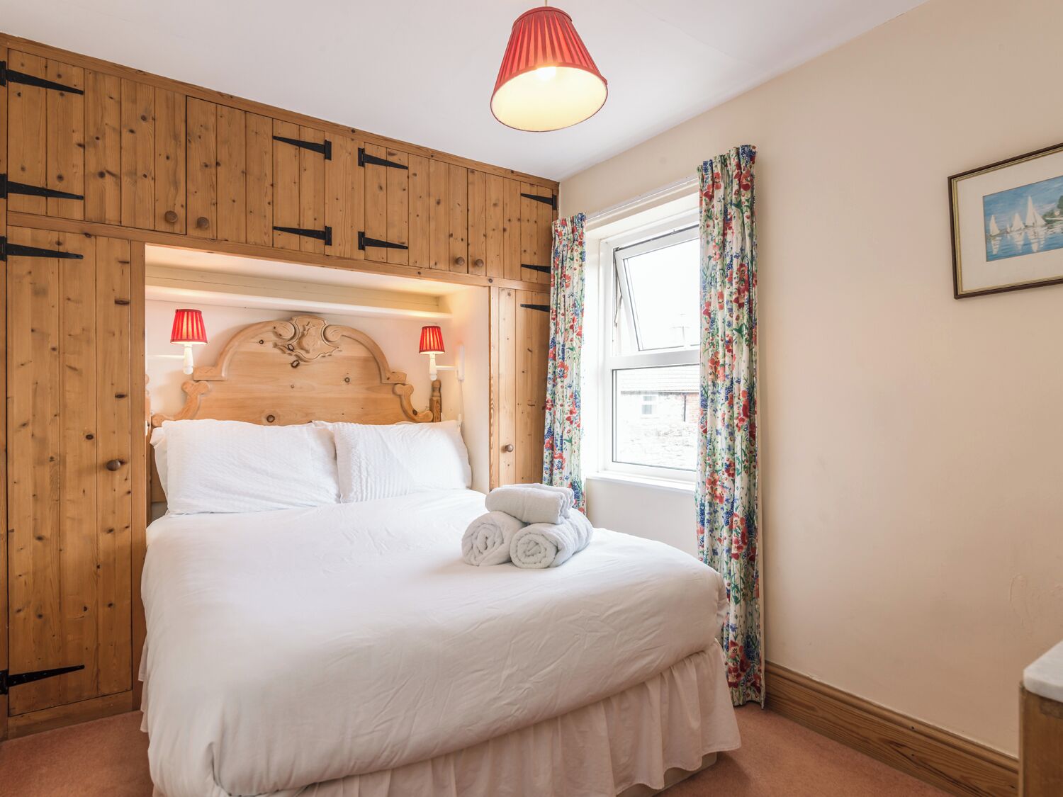 Une chambre à coucher avec une tête de lit en bois, un grand lit recouvert de draps blancs, deux lampes de chevet et une fenêtre ornée de rideaux à motifs floraux.