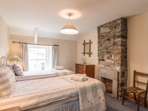 A bedroom with two beds, a stone fireplace, a wooden dresser, and a chair. - Fisherman's Cottage (Seahouses)