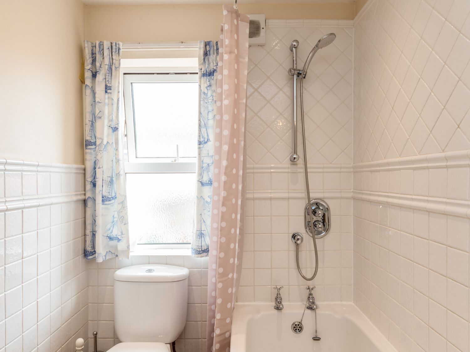 A bathroom with a toilet, a shower with a handheld showerhead, and a window with curtains.