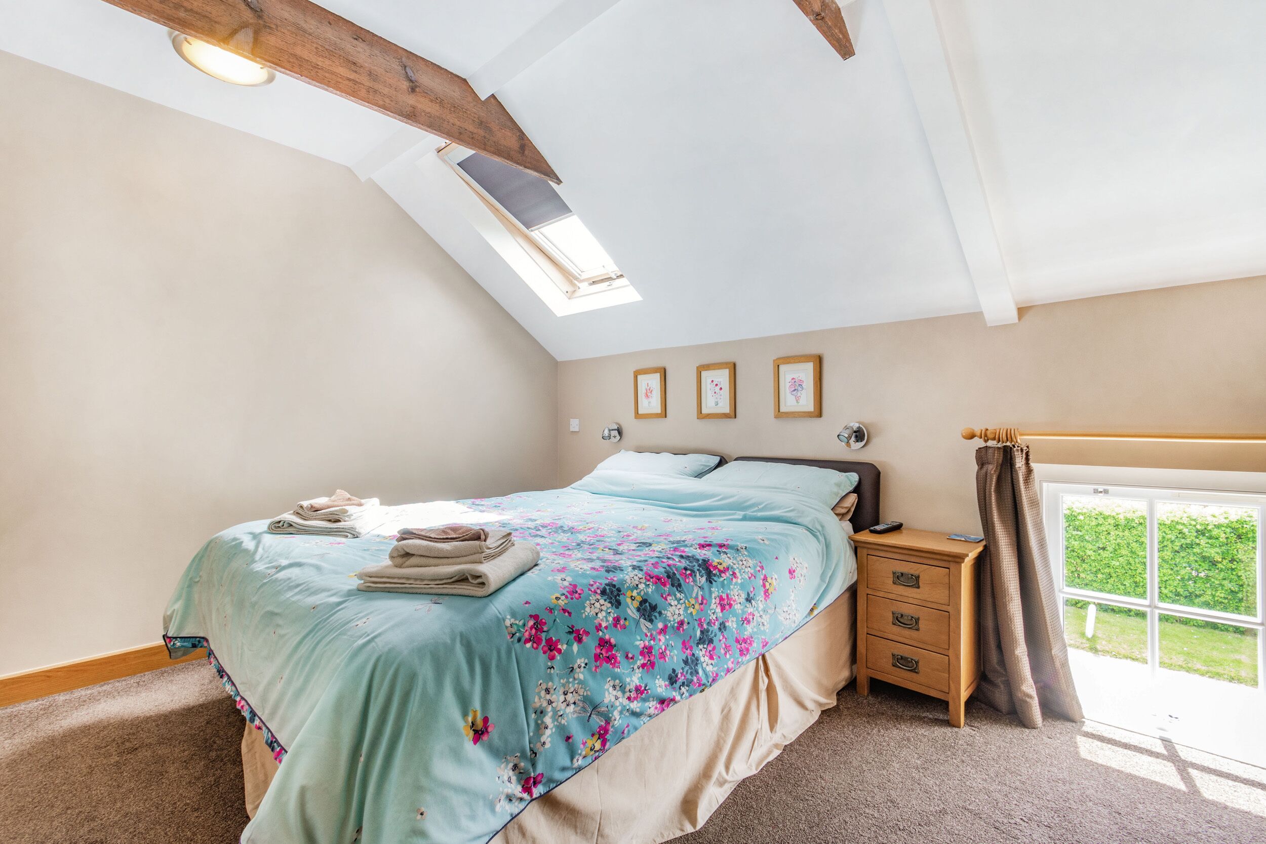 A bedroom with a skylight, a bed with a floral bedspread, a nightstand, and a window with a view of greenery.