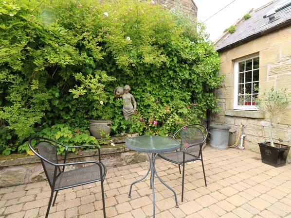 Une terrasse avec une table et des chaises, entourée d’une végétation luxuriante et d’un mur de pierre.