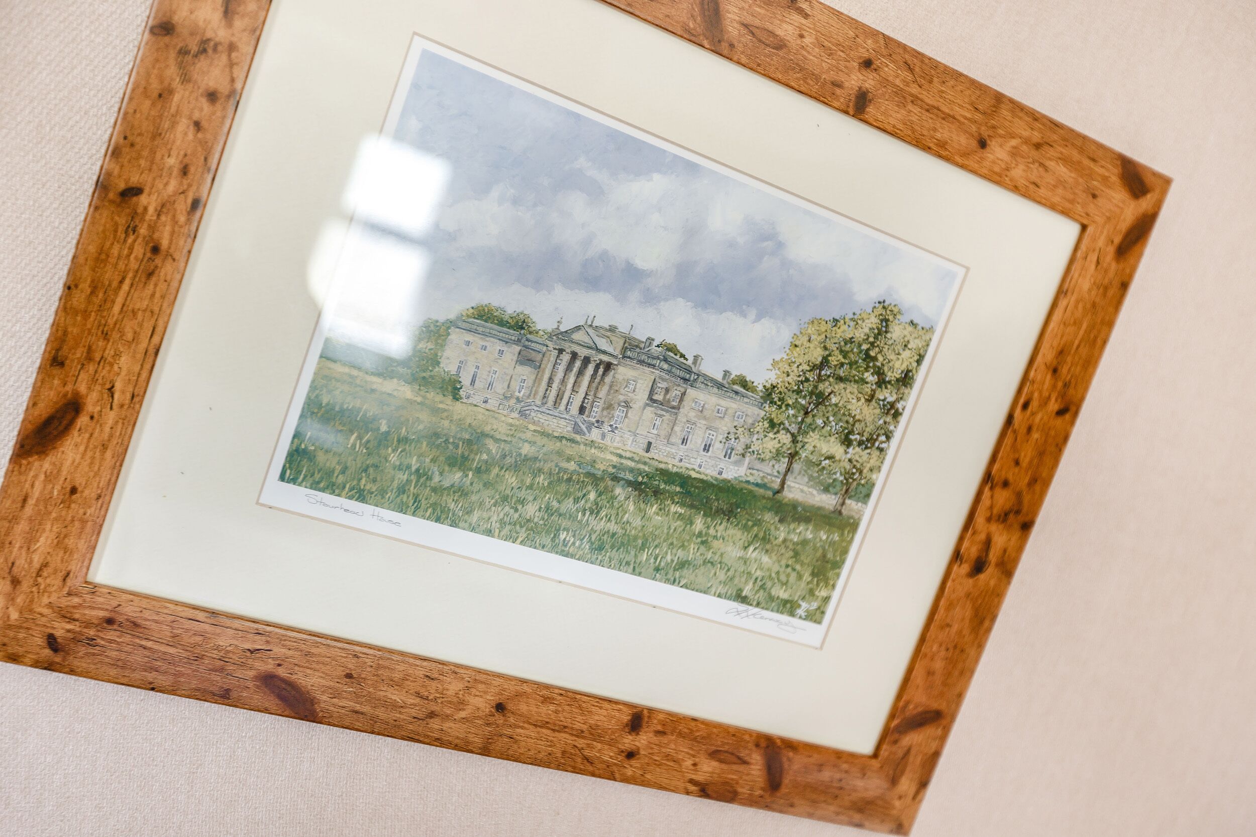 Une œuvre d’art encadrée représentant un bâtiment classique avec des colonnes, un arbre et un premier plan verdoyant.