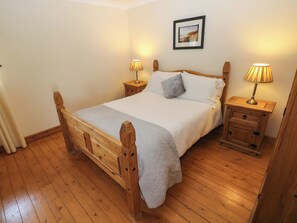 A wooden four-poster bed with a white bedspread and pillows, flanked by nightstands with lamps, in a room with wooden flooring and a framed picture on the wall.
